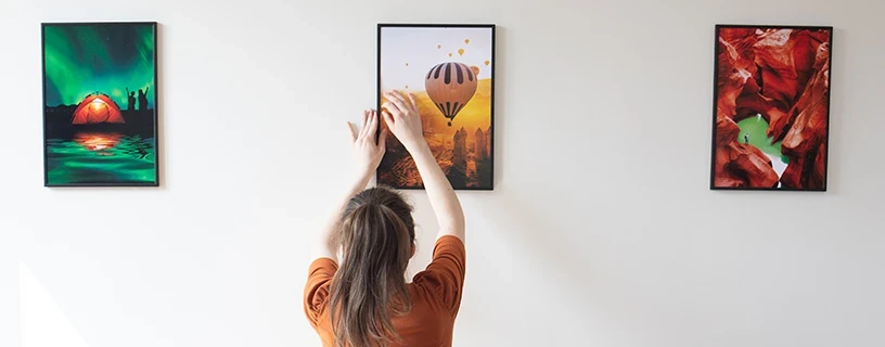 Junge Frau hängt Bilderrahmen an weißer Wand auf, darin Farbdrucke mit Pro-Design